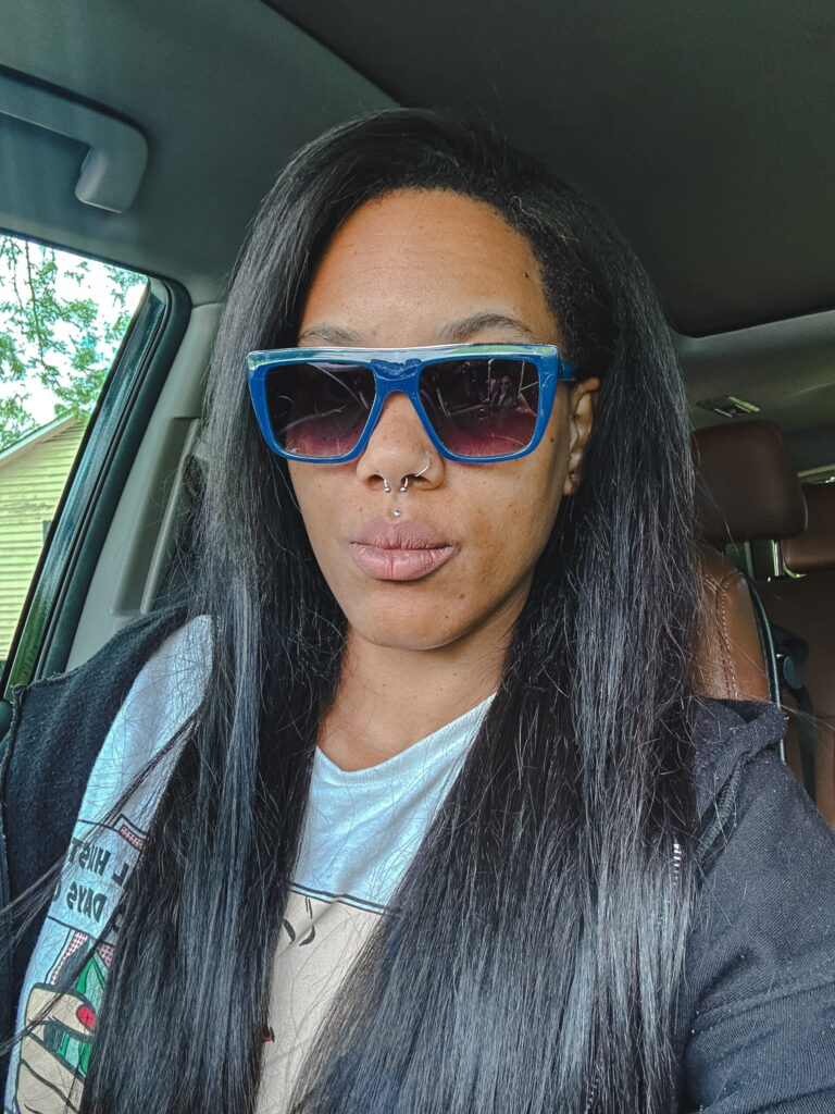 girl with long black hair sitting in her car wearing Sojos blue green flat top sunglasses